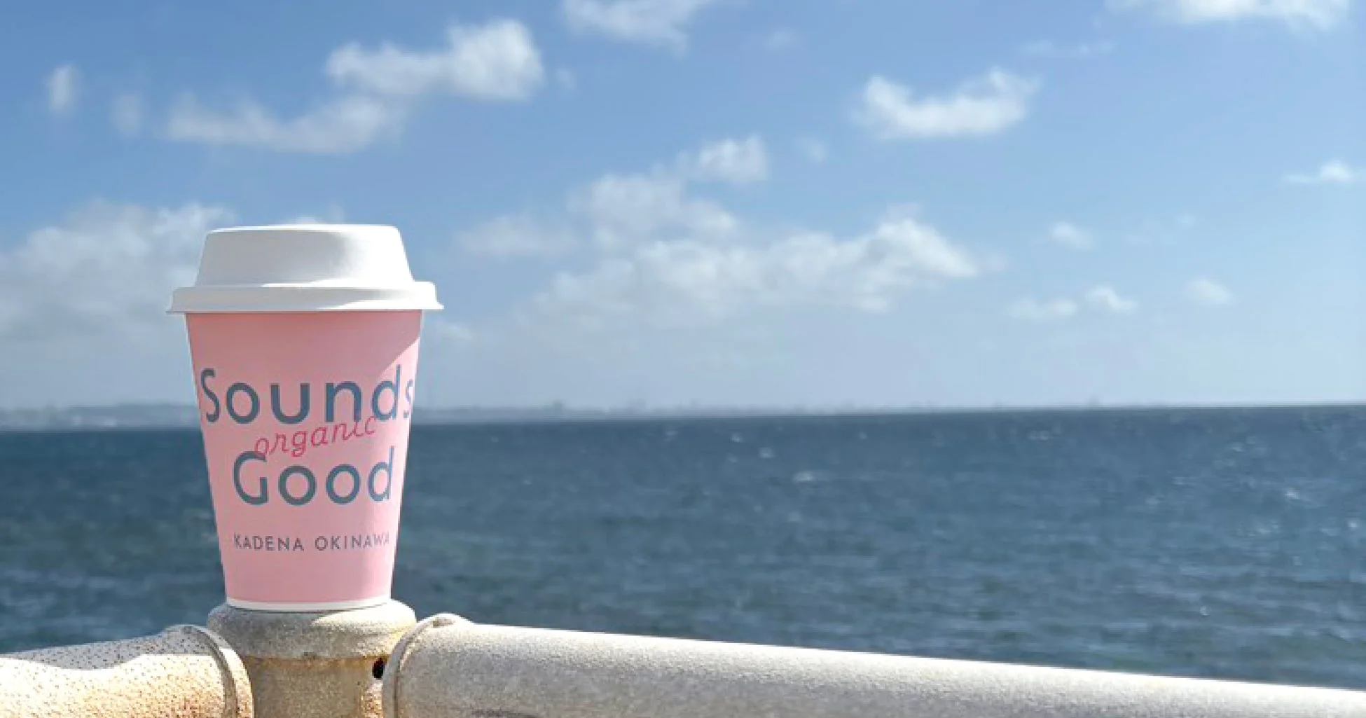 海を背景に、ピンク色のコーヒーカップが手すりの上に置かれています。青い空と広がる海が開放的な景色を作り出し、リラックスした雰囲気を醸し出しています。